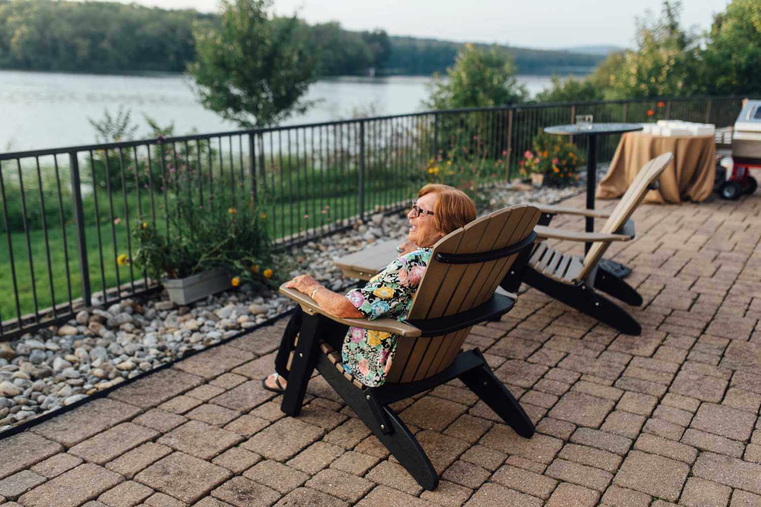 A woman sits in an adirondack chair on a patio at the lake house inn, overlooking a lake.