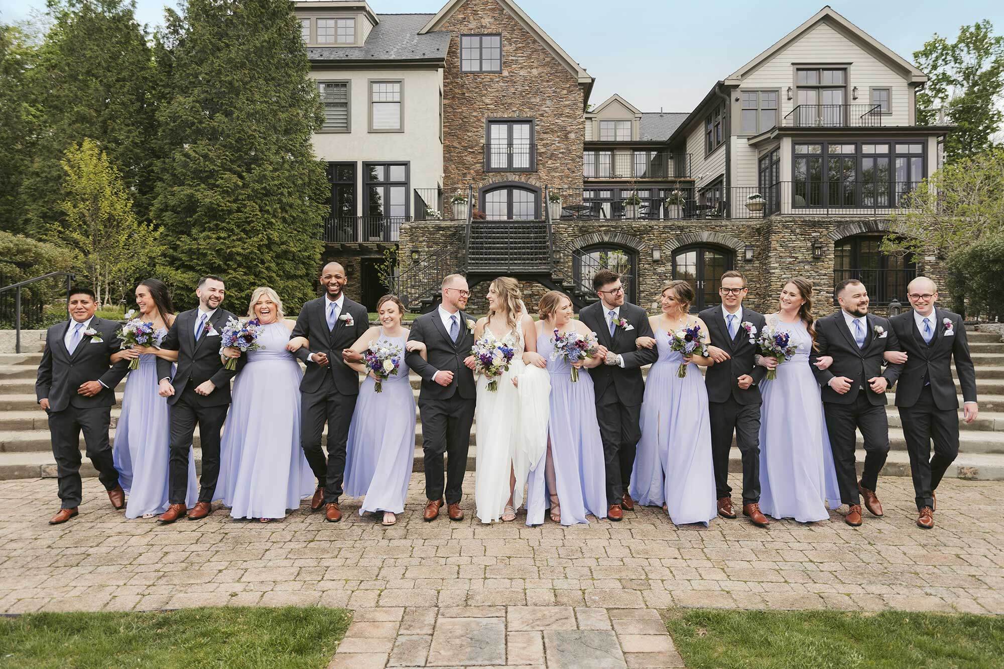 A group of bridesmaids and groomsmen pose in front of a mansion.