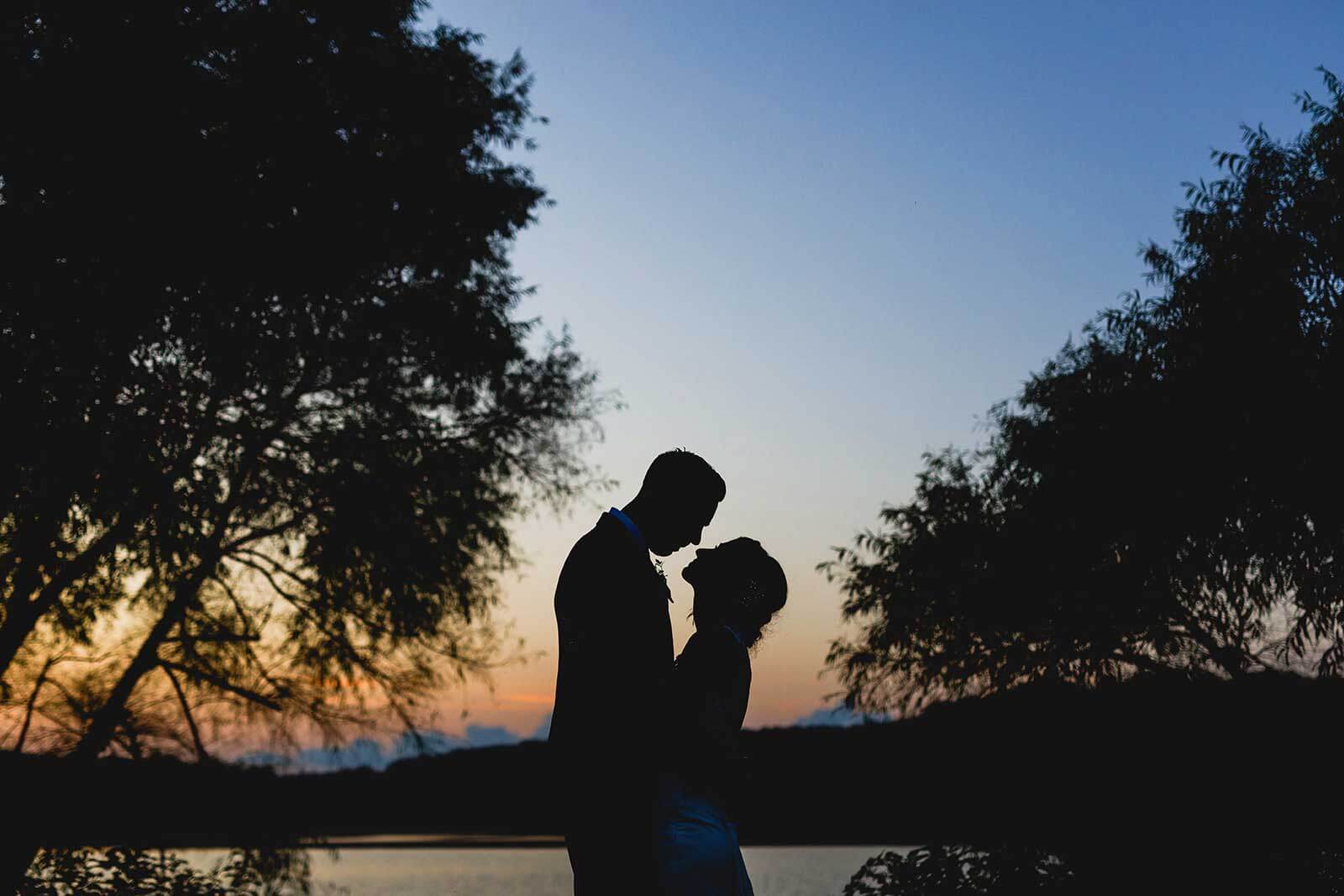 A bride and groom are silhouetted by a lake at sunset, capturing a memorable wedding experience.