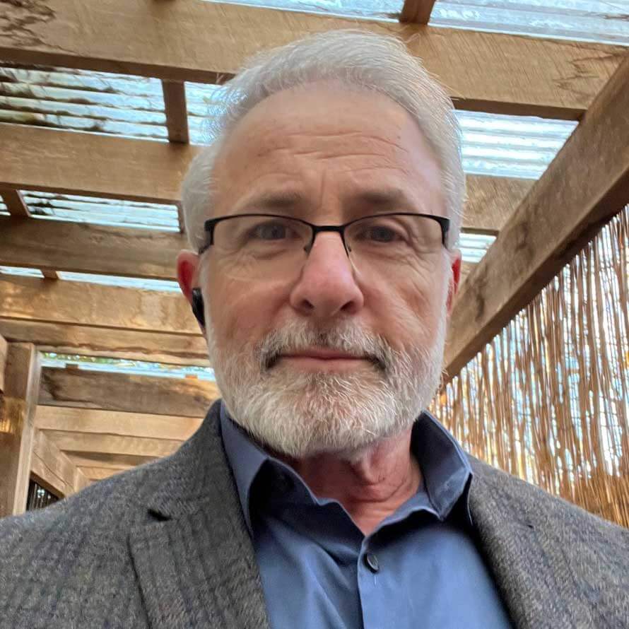 A man with glasses and a beard is standing in front of a wooden structure.