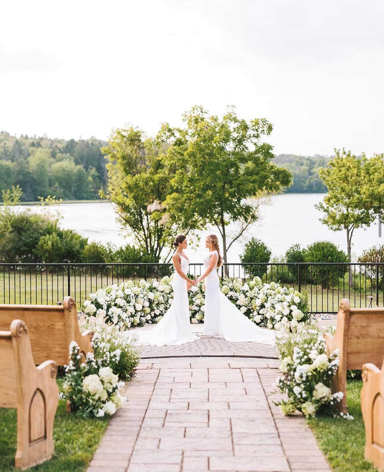 Two brides standing in front of a bench in front of a lake.