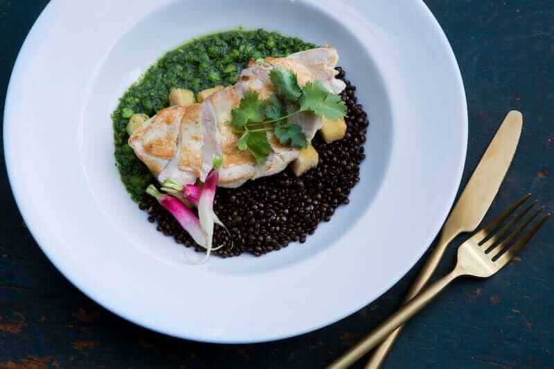 Grilled chicken breast served on a bed of green sauce with a side of black lentils, garnished with herbs and radishes, accompanied by golden cutlery.