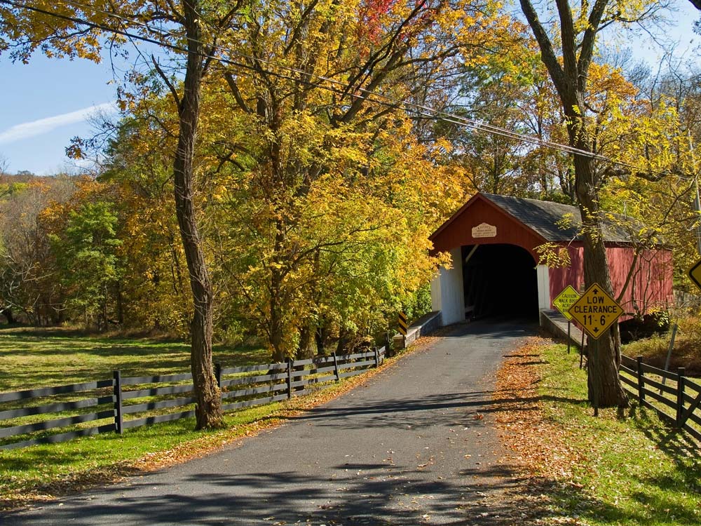 Autumn scene with a red covered bridge over a road, surrounded by colorful trees and a white fence near the lake house inn, and a clearance sign on the right.