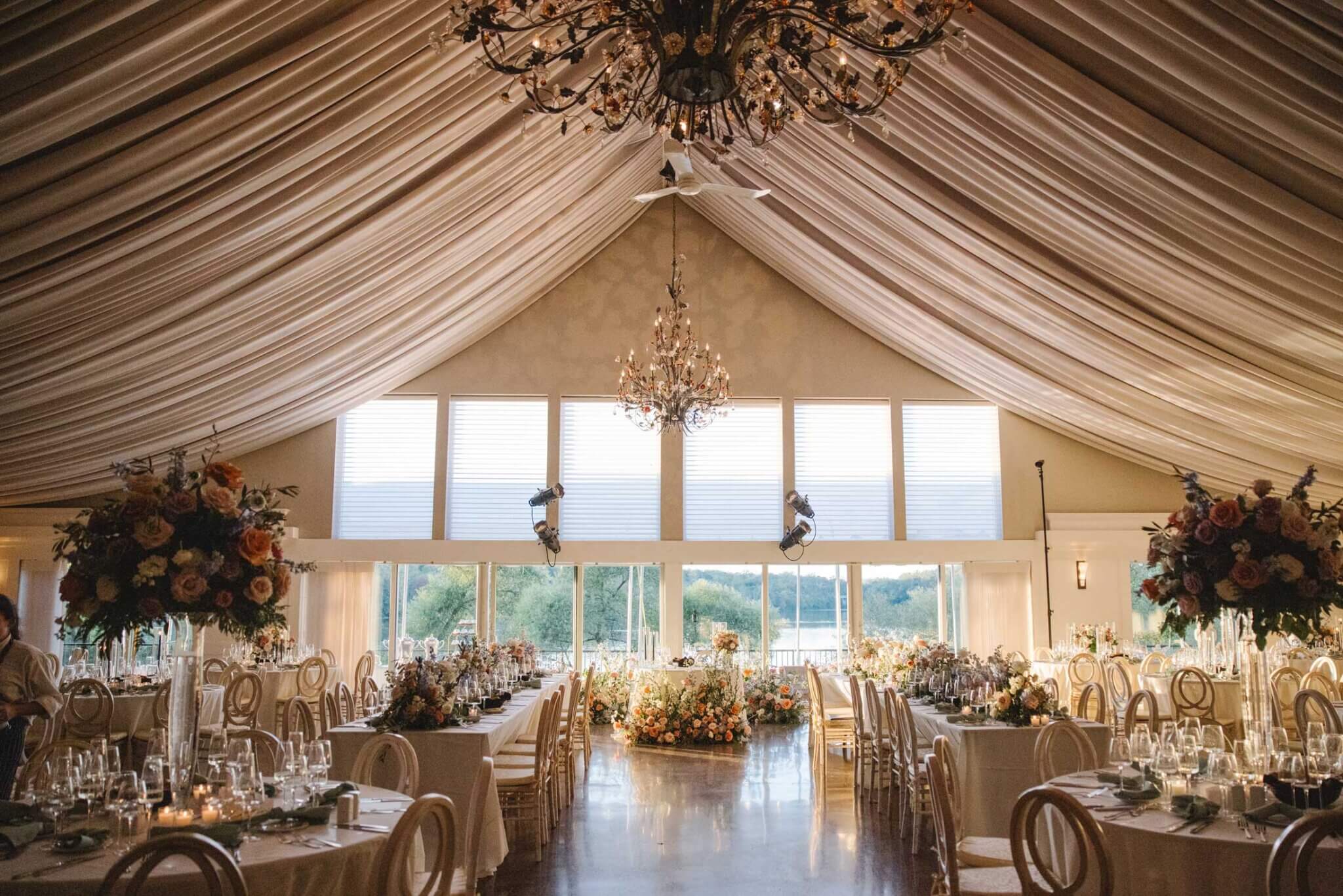 Elegant banquet hall with draped ceiling, chandeliers, large windows, round tables set for dining, and floral centerpieces; natural light fills the room.