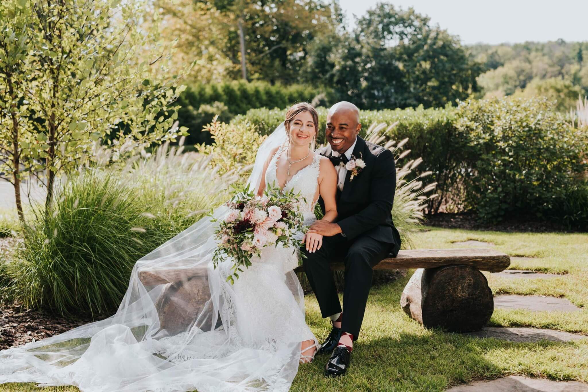 A bride in a white dress and veil sits next to a groom in a black suit on a wooden bench outdoors, smiling and holding a bouquet of flowers, capturing the joy of their unforgettable wedding experience.