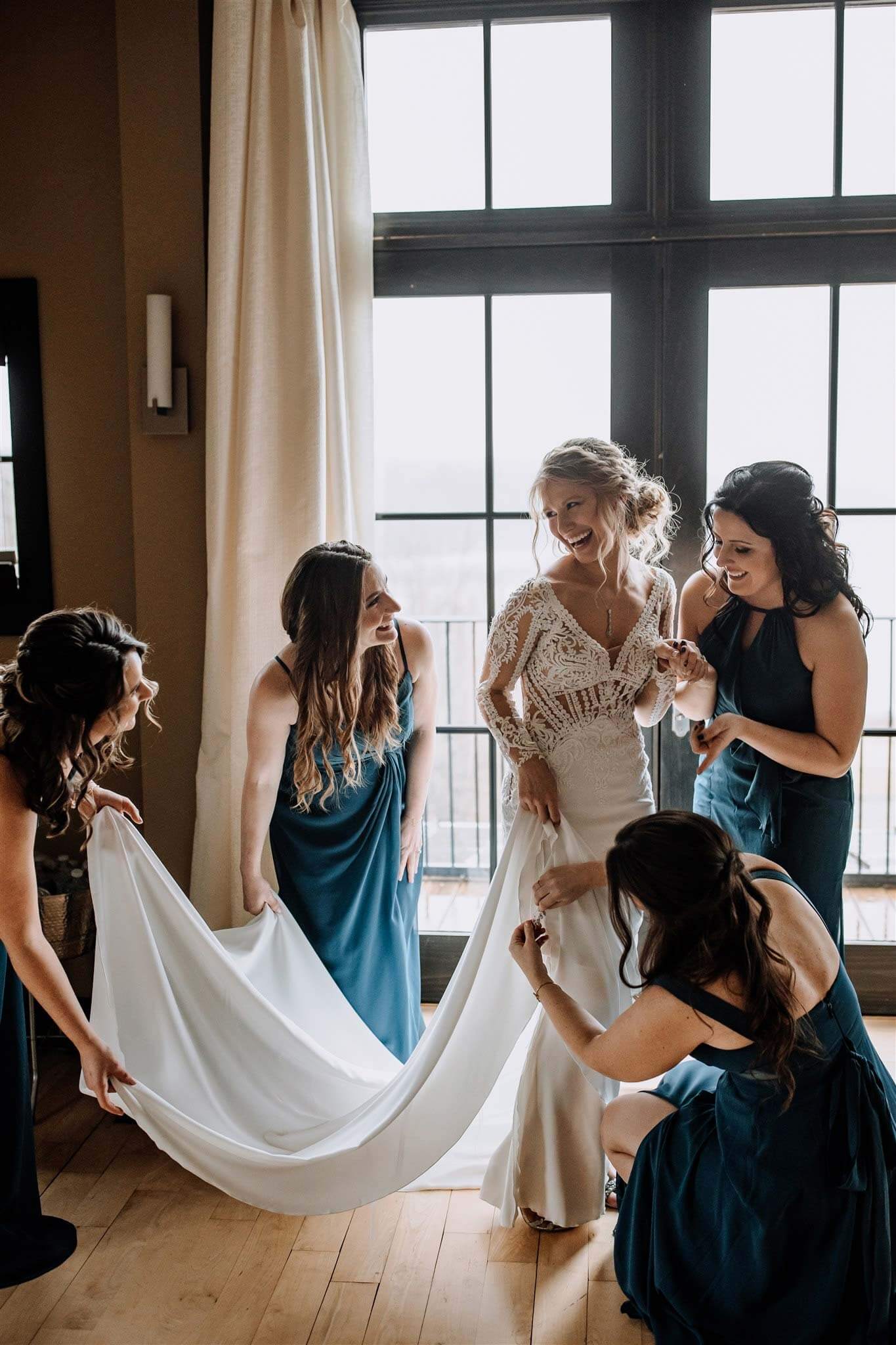 Four women in blue dresses help a bride adjust her white gown in a sunlit room with large windows, capturing a joyful wedding experience.