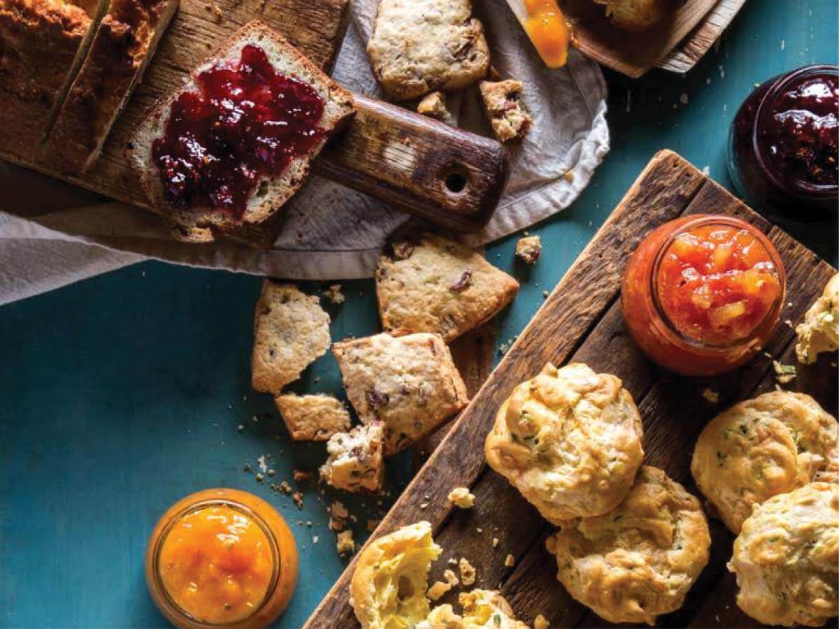 Assorted breads and biscuits on wooden boards, served with jars of orange and red jam, create a charming wedding experience, with crumbs scattered on a blue tabletop.