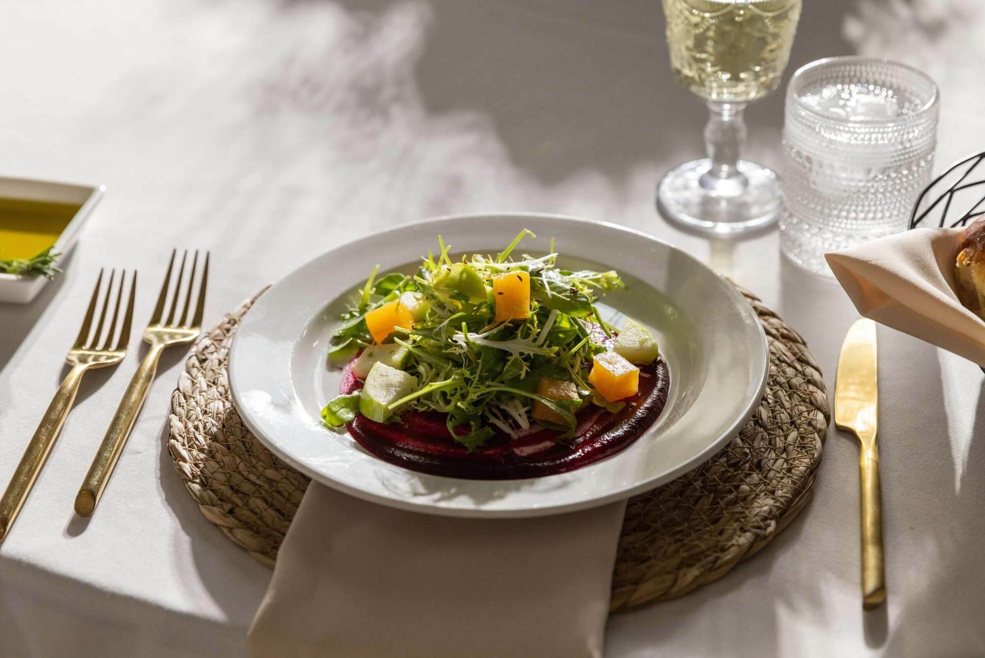A white plate with a green salad and diced melon sits on a woven placemat with gold cutlery, a bread roll, and a glass of white wine—perfect for an elegant wedding experience.