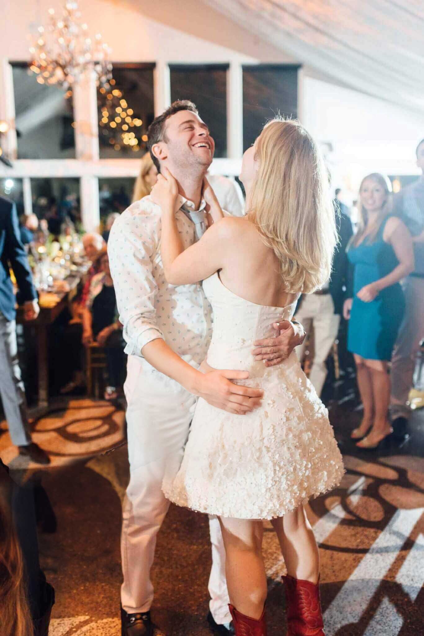 lake-house-inn-rehearsal-dinner-retouch A couple dances closely at an indoor event; the woman wears a white strapless dress and red boots, and the man is in a light shirt and pants. Guests watch in the background.