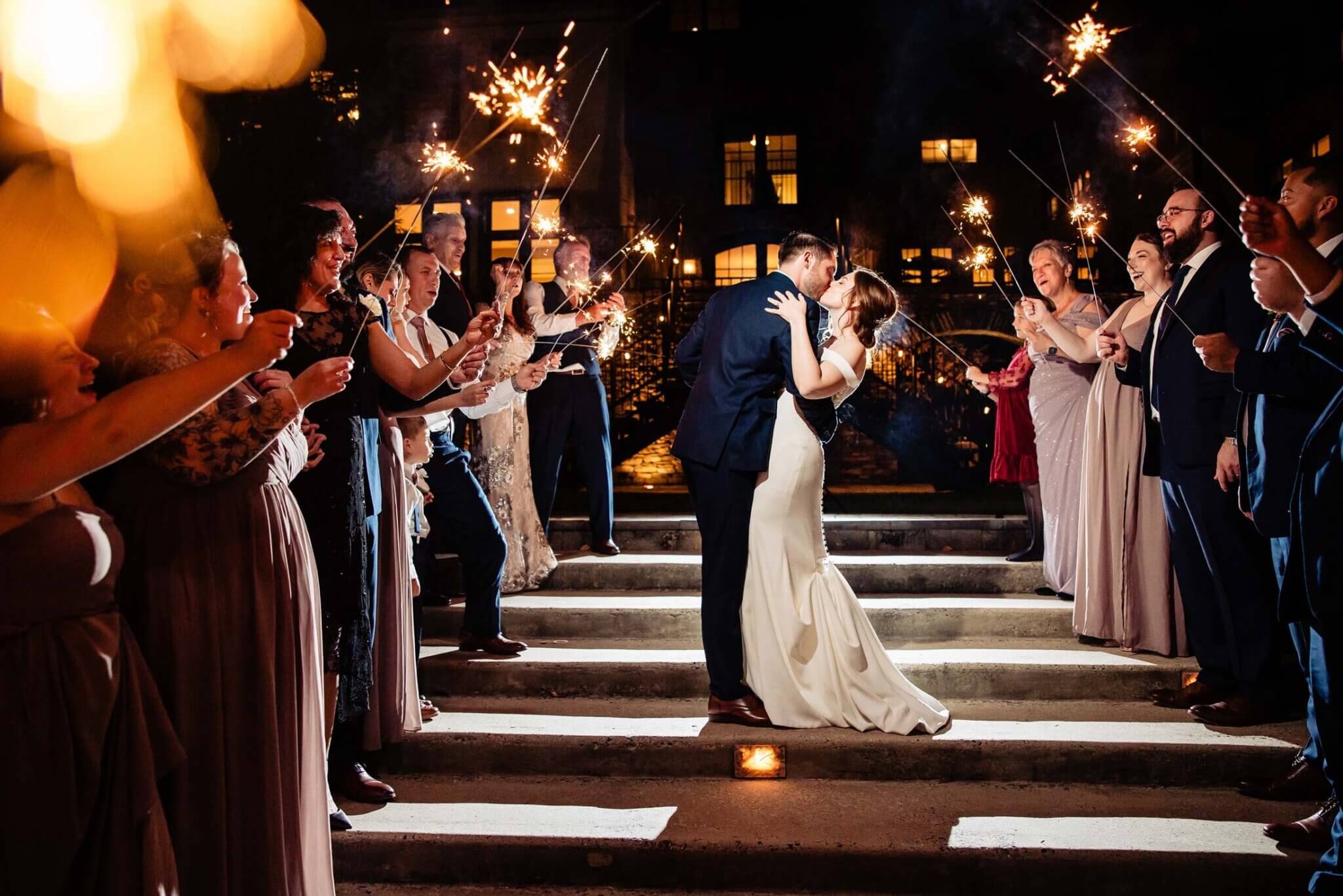 A bride and groom kiss on outdoor steps at night, surrounded by guests holding sparklers and standing in two lines, creating a magical wedding experience.