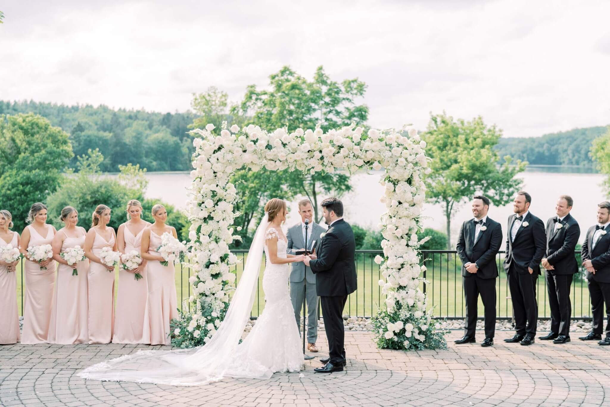 A bride and groom exchange vows under a white floral arch outdoors, surrounded by bridesmaids in light dresses and groomsmen in dark suits, creating an unforgettable wedding experience.