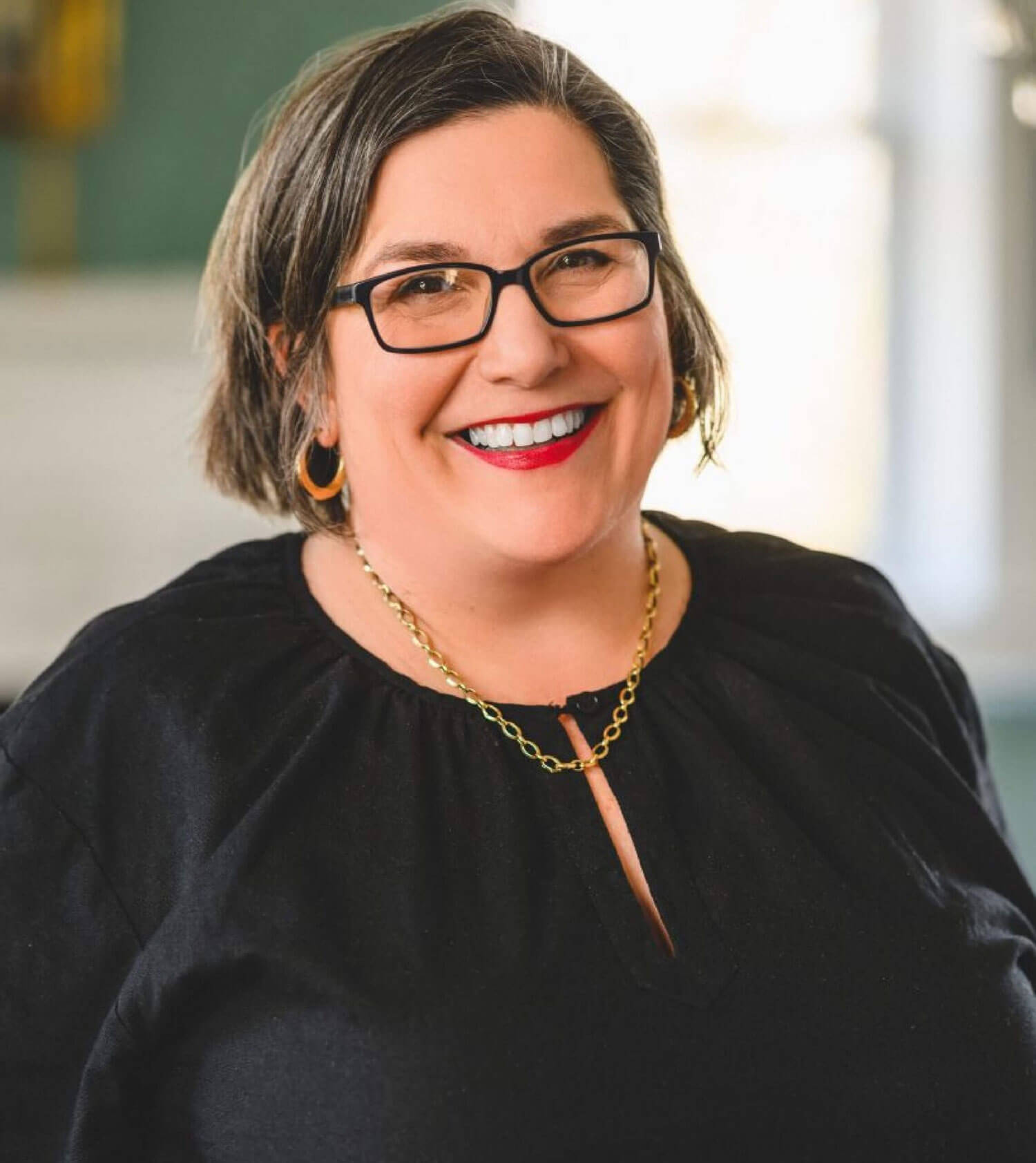 A woman with short brown hair, glasses, and red lipstick smiles while wearing a black top, gold hoop earrings, and a gold chain necklace.