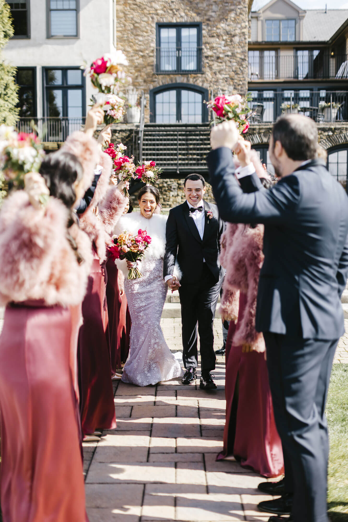 A bride and groom walk hand in hand outdoors, smiling, as their wedding party in burgundy attire and pink fur shawls cheer and raise bouquets on either side of the path.