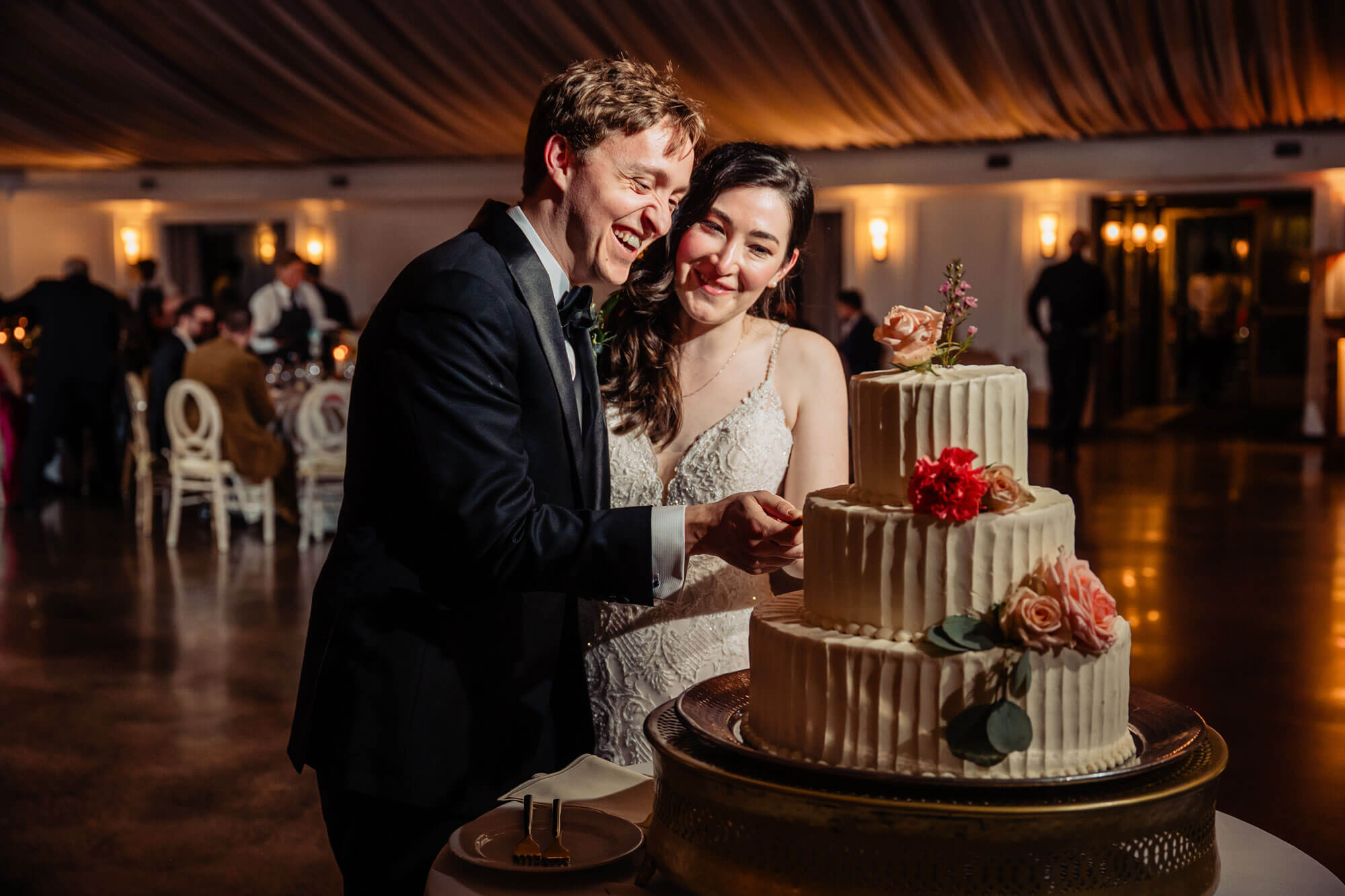 A bride and groom, both smiling, cut a tiered wedding cake decorated with flowers at their reception, showcasing one of our elegant wedding packages.