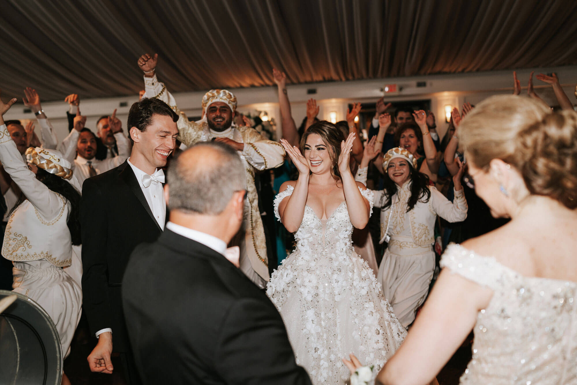 A bride and groom smile and clap while surrounded by guests dancing and celebrating at a wedding reception under a draped ceiling.