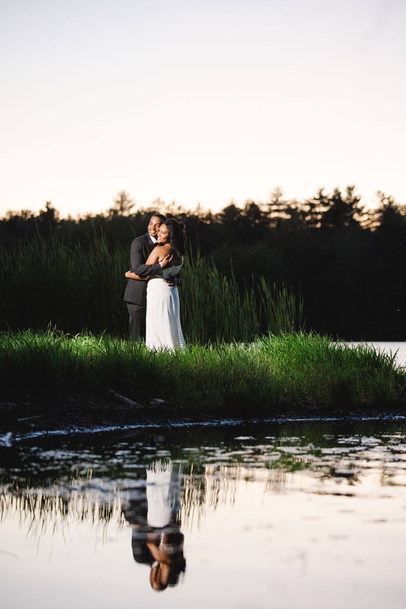 A couple embraces on a grassy lakeshore at dusk, with their reflection visible in the calm water in the foreground.