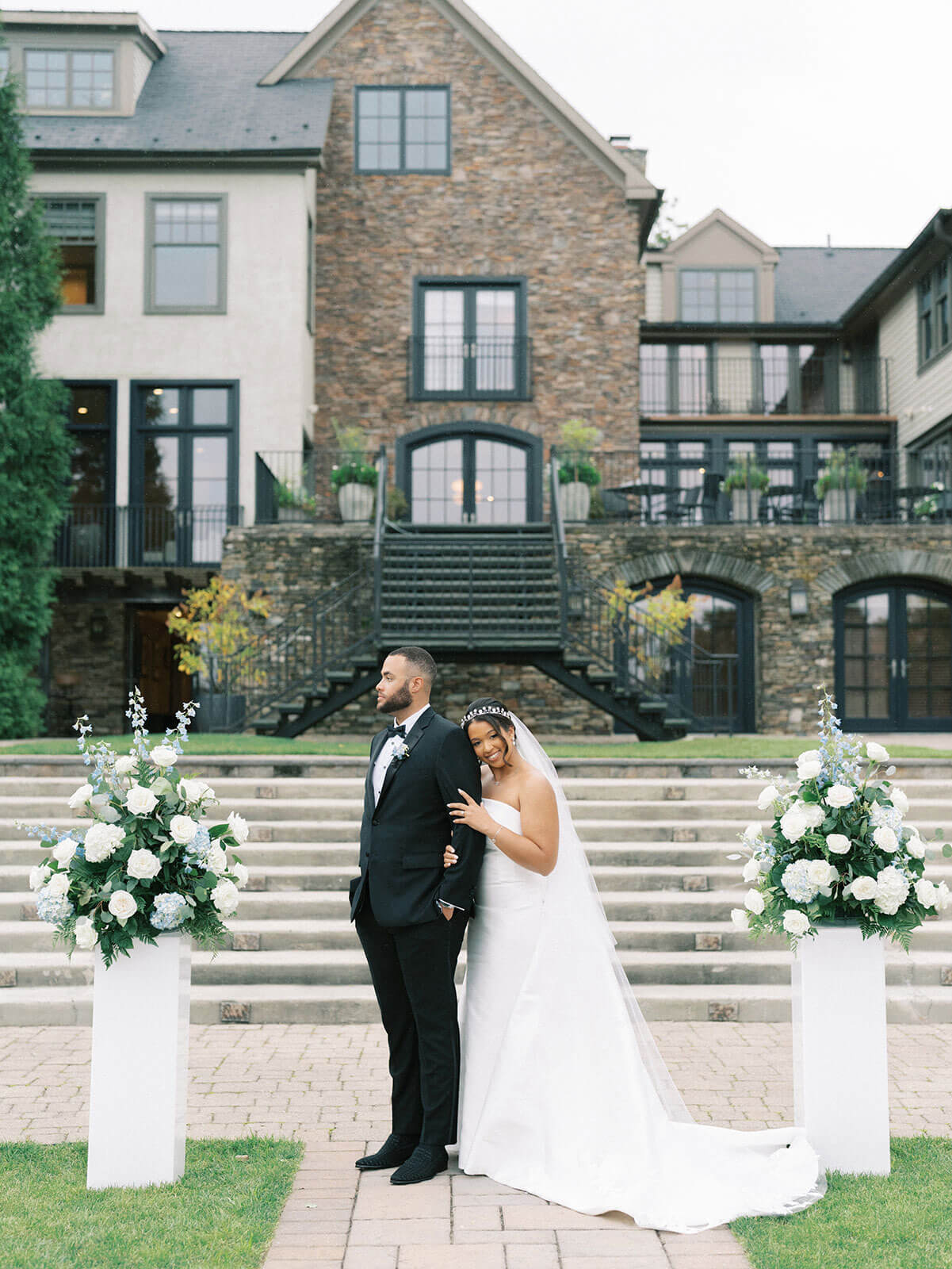 A bride in a white gown and veil stands behind and holds her groom in a black suit, capturing a memorable wedding experience outdoors in front of a large house with stone steps and floral arrangements.