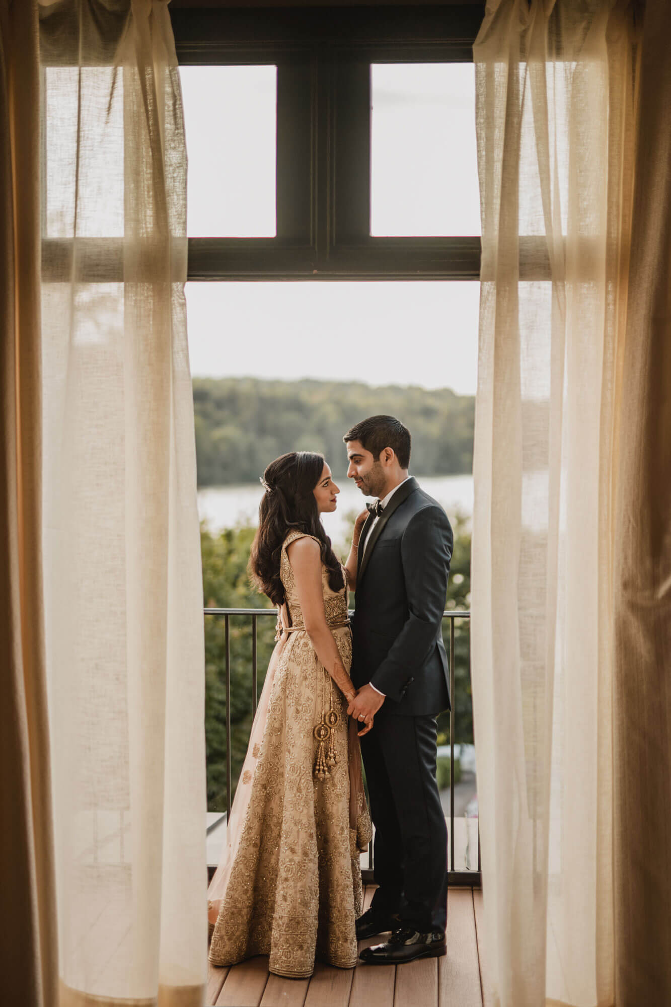 A couple dressed in formal attire stands facing each other on a balcony, holding hands, framed by open curtains with a scenic outdoor view in the background.
