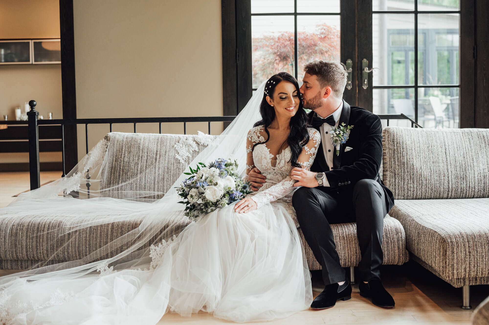 A bride and groom sit together on a couch indoors, with the groom kissing the bride on the temple. The bride holds a bouquet and wears a long veil and lace dress.