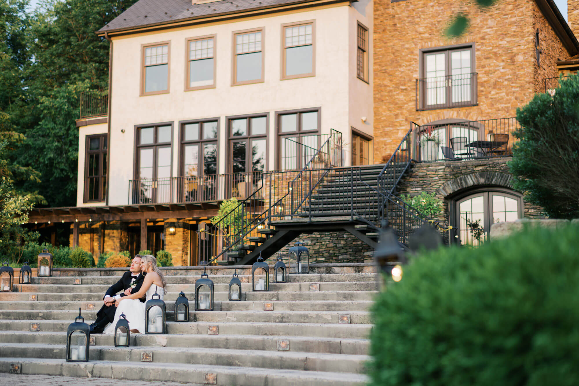 A couple in formal attire sits on outdoor stone steps surrounded by lanterns in front of a large house with many windows and a black-railed staircase.