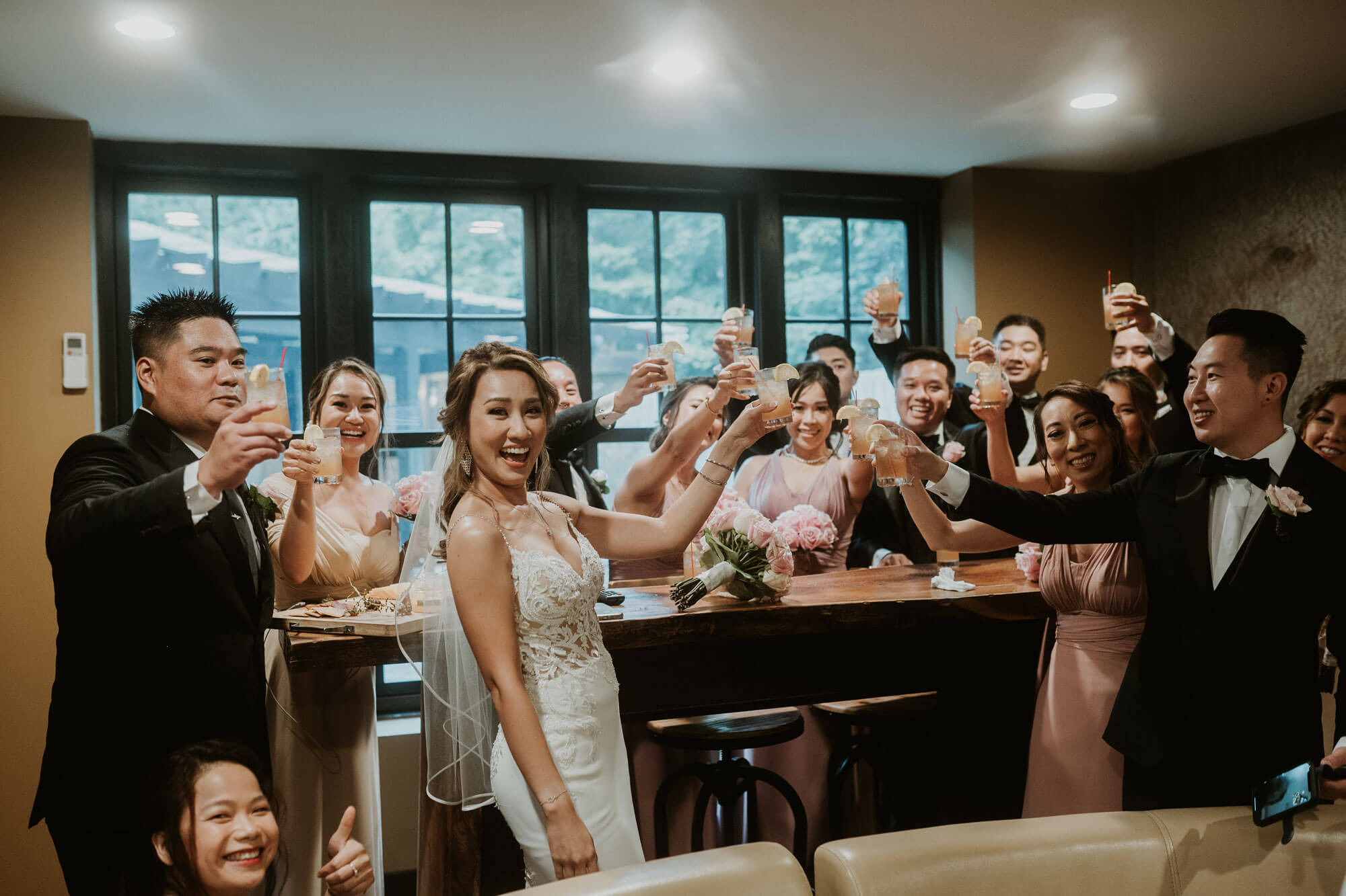 A bride and groom staying at the lake house inn toast with their wedding party in a room, smiling and raising glasses, with guests gathered around a wooden table.