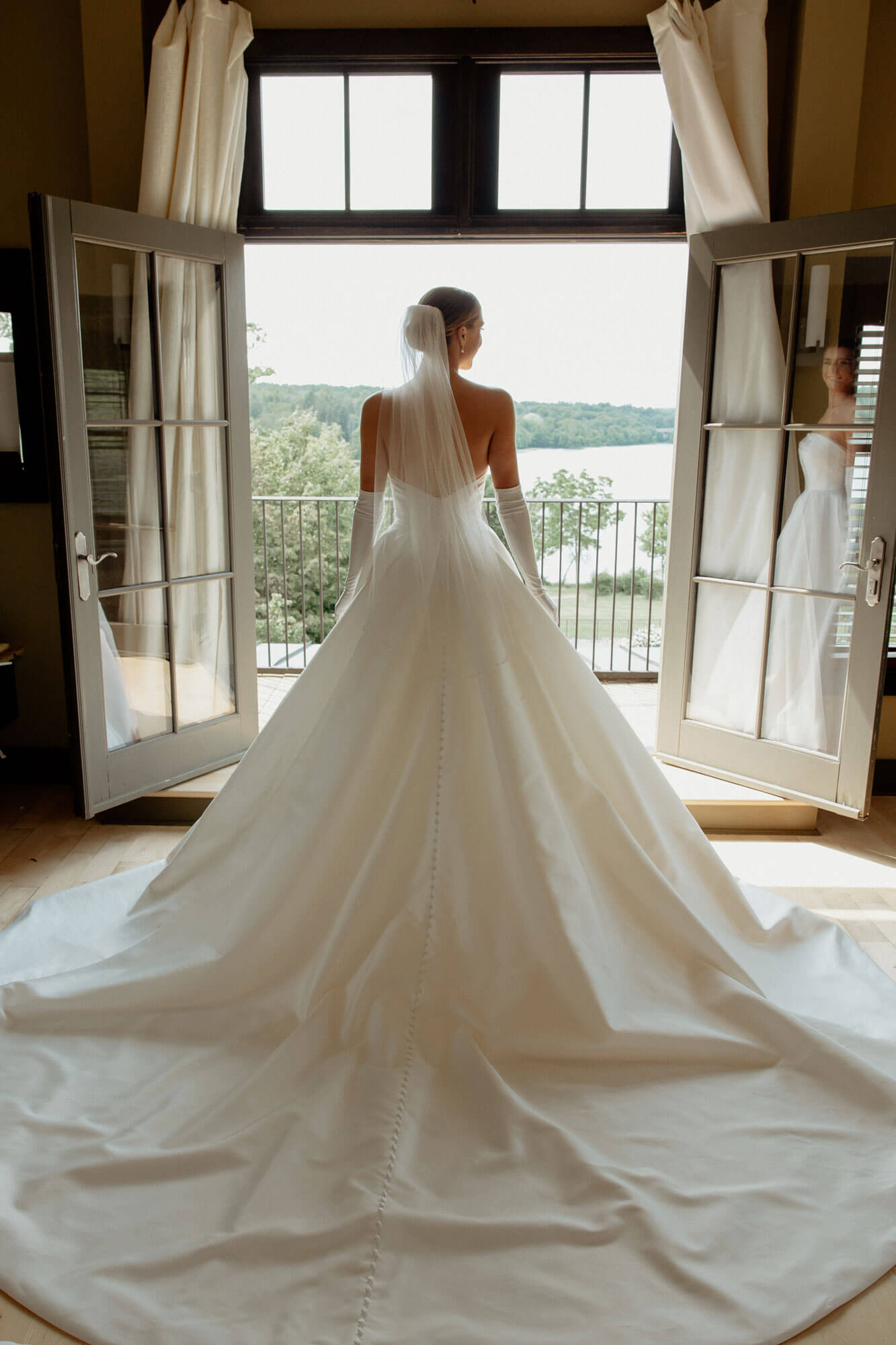A bride in a long white wedding gown and veil stands facing open double doors, looking out over a balcony with trees and a body of water in the background.