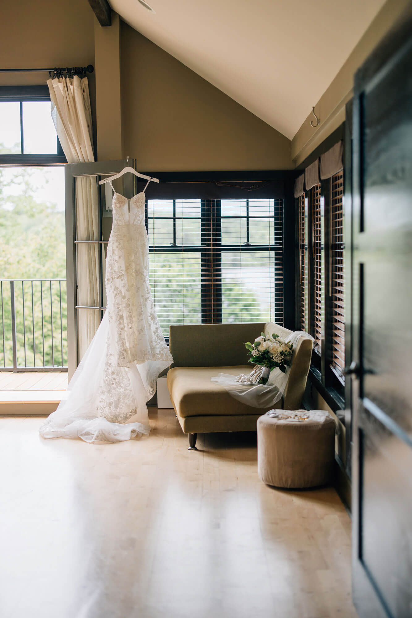 A white lace wedding dress hangs by a window near a beige chaise lounge with a bouquet of flowers and a round ottoman in a sunlit room.