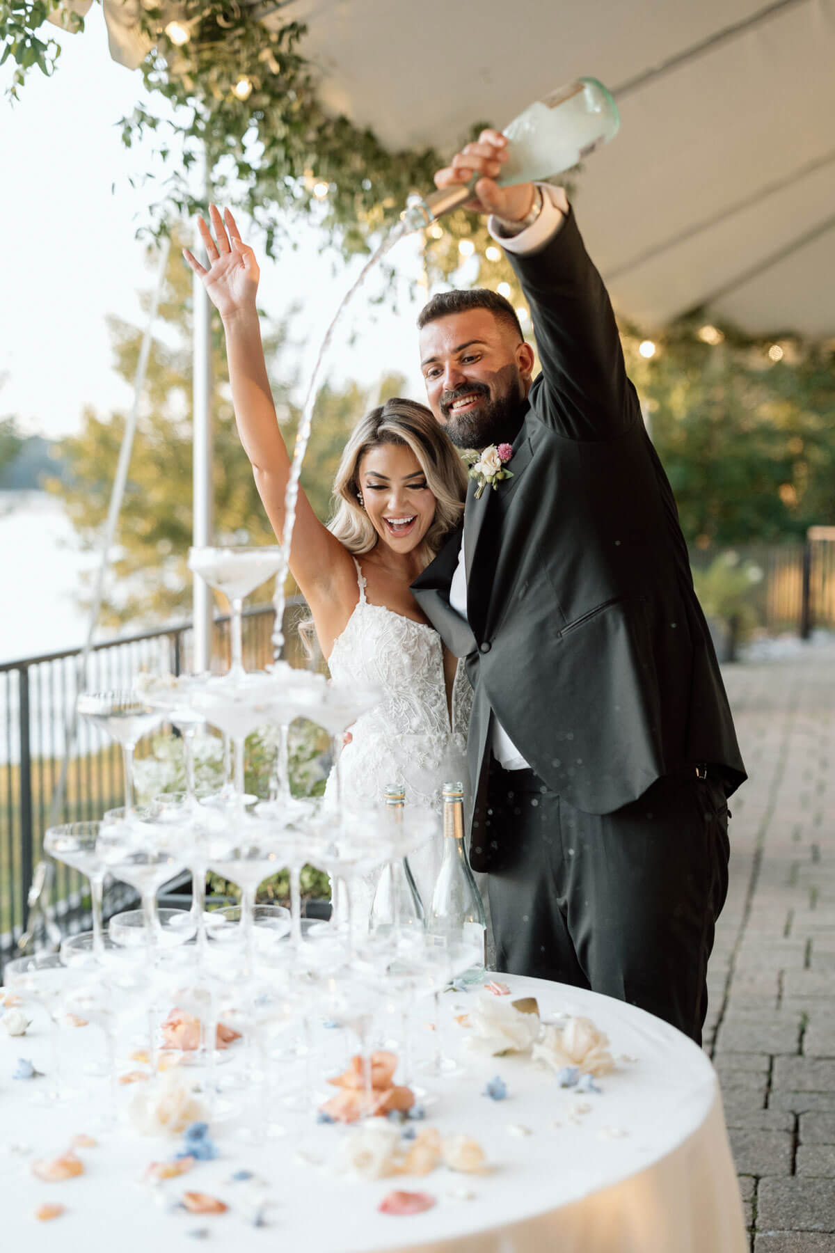 A bride and groom smiling and pouring champagne into a tower of glasses at an outdoor wedding reception, creating a magical Wedding Experience to remember.