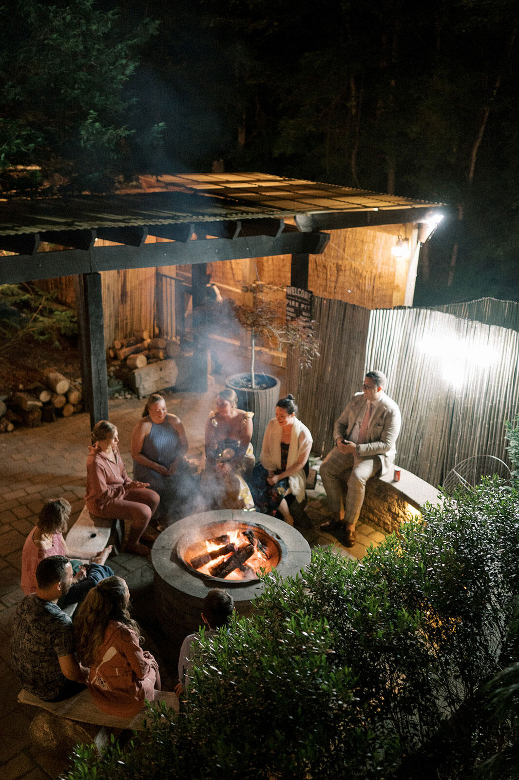 A group of people sit around a fire pit at night in an outdoor patio area, talking and relaxing under a wooden pergola with greenery and trees nearby while staying at the lake house inn.