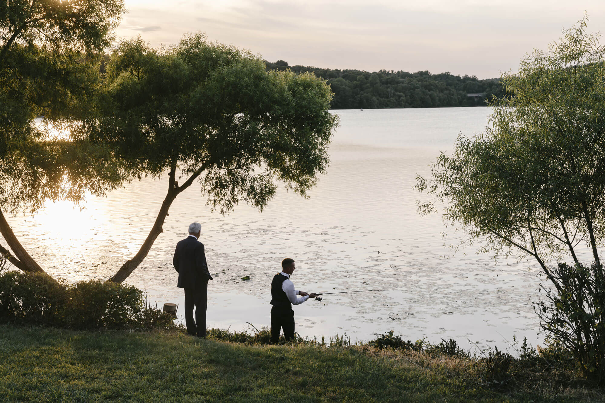 Two people dressed in formal clothes stand by a lakeshore under trees, with one person fishing as the sun sets over the water.