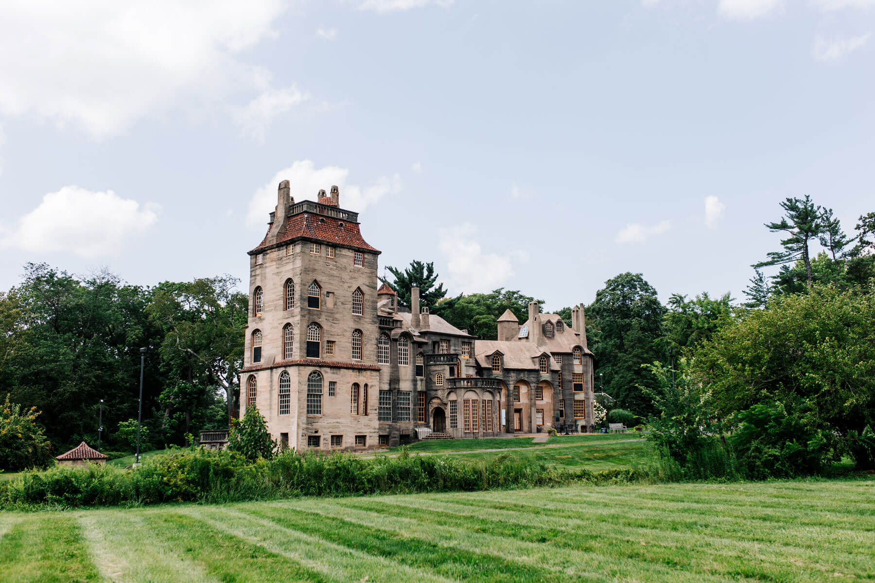 A large, historic concrete mansion with multiple levels and towers sits surrounded by green grass and trees under a partly cloudy sky—perfect for staying at the Lake House Inn.