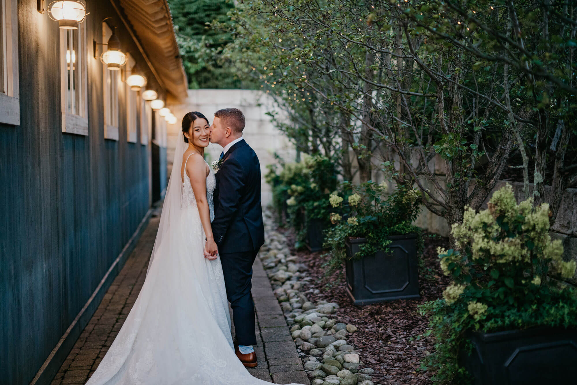 A bride and groom stand outside on a stone path, holding hands and smiling, with greenery and string lights in the background, capturing a magical wedding experience.