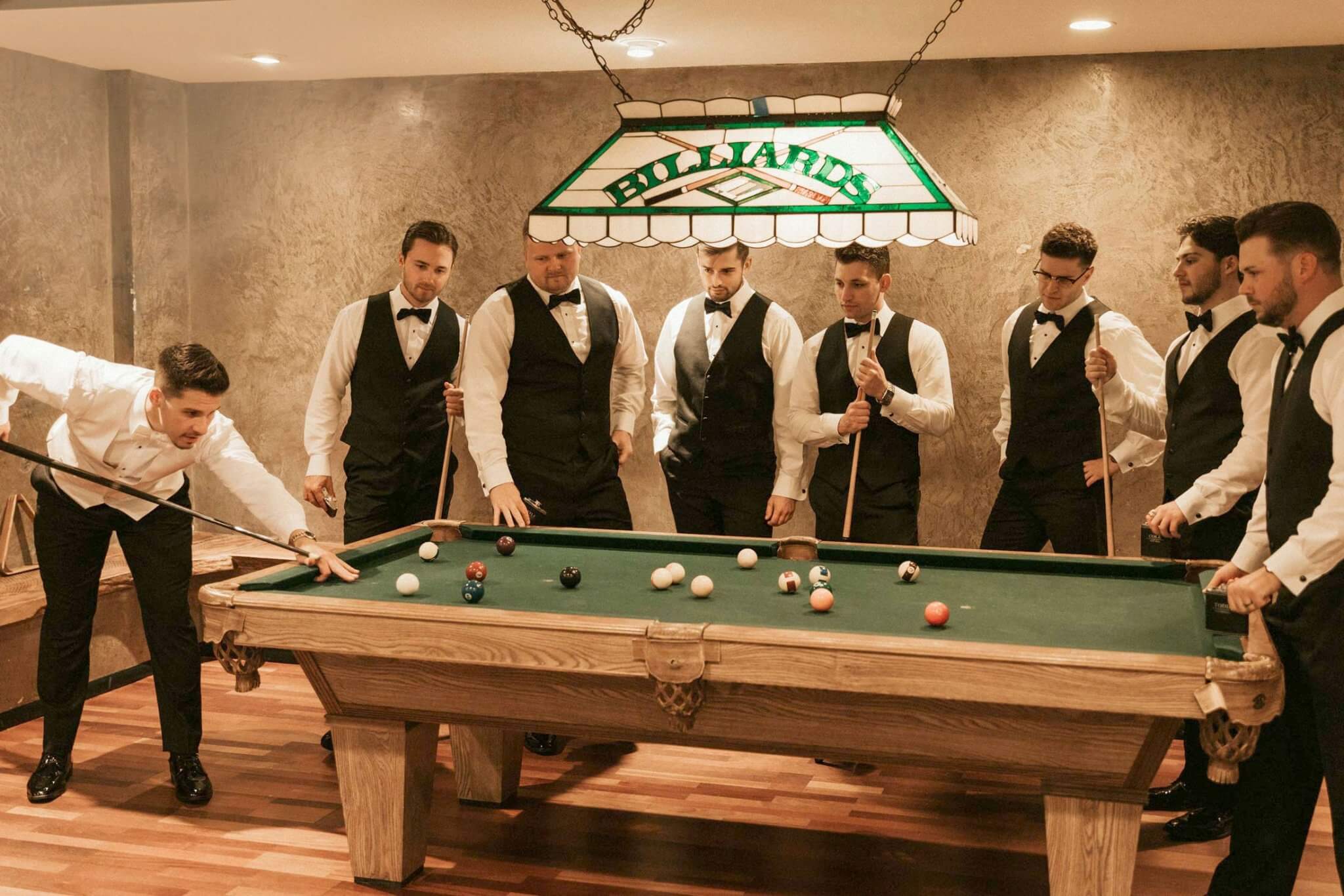 Eight men in formal attire gather around a billiards table, sharing laughter and anticipation as one takes a shot—capturing a memorable Wedding Experience beneath the glowing billiards sign.