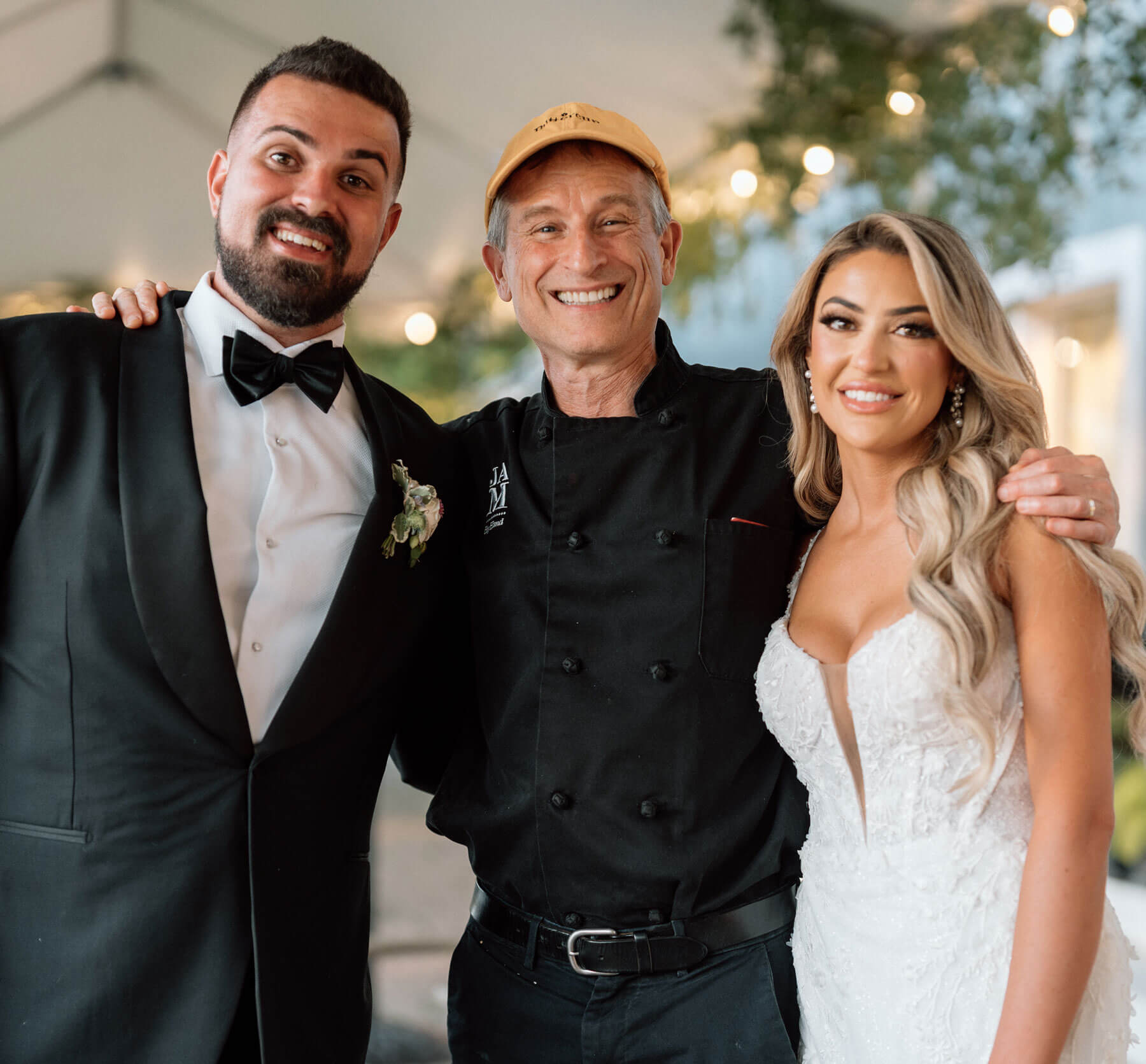A man in a tuxedo, a chef in a black uniform and yellow cap, and a woman in a white wedding dress stand together smiling under a canopy.
