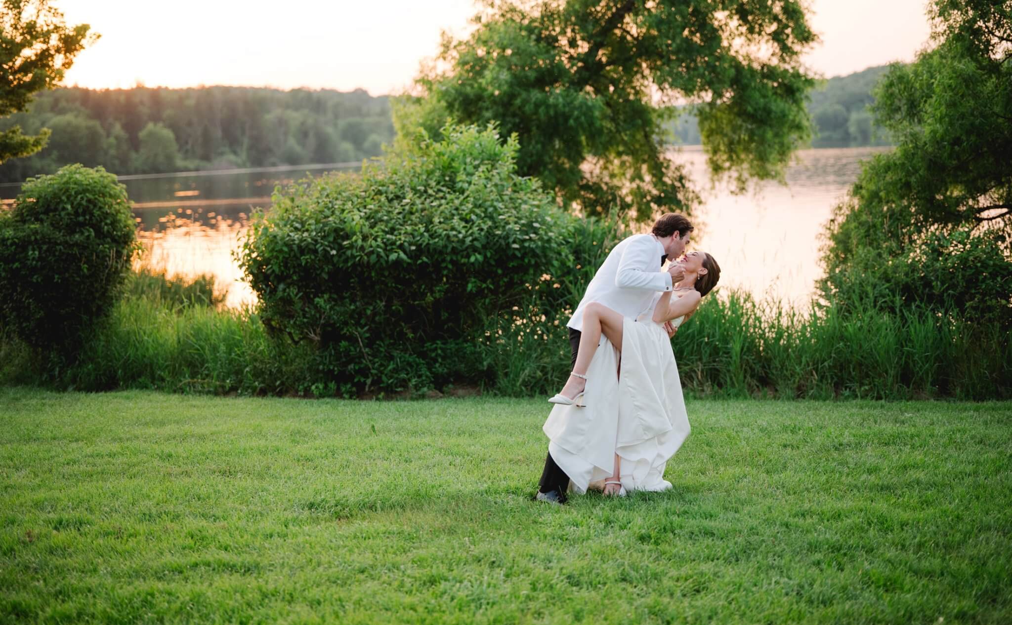 A groom in a white suit lifts and kisses a bride in a white dress on a grassy lawn near a lake at sunset.