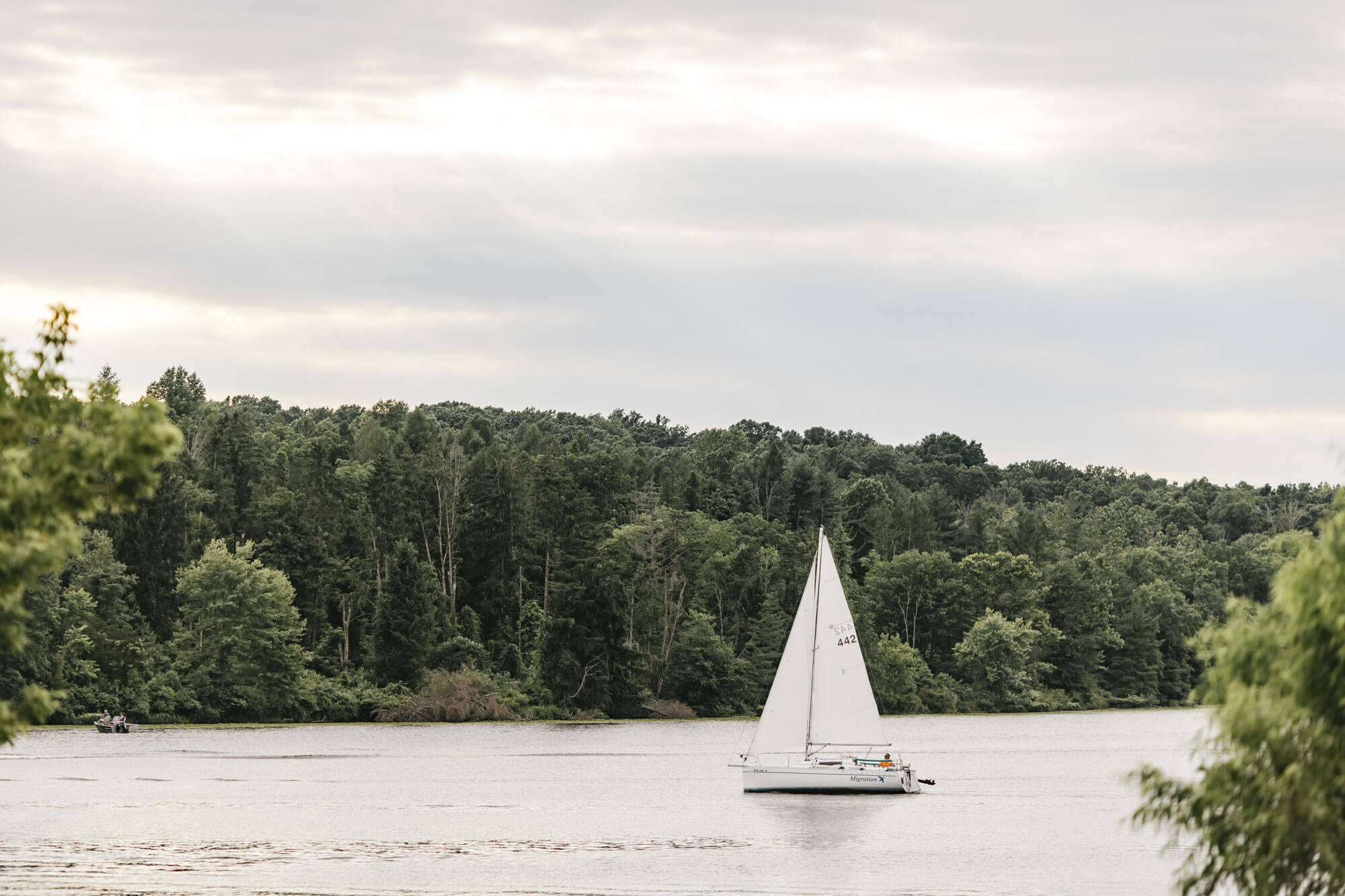 A white sailboat moves across a calm lake, surrounded by dense green trees under a mostly cloudy sky.