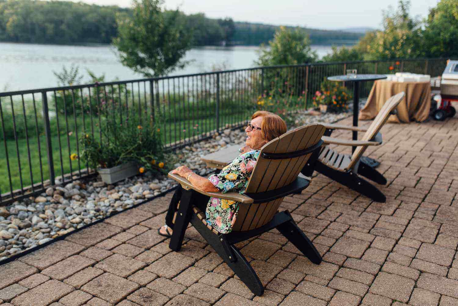 A woman sits in an adirondack chair on a patio at the Lake House Inn, overlooking a lake.