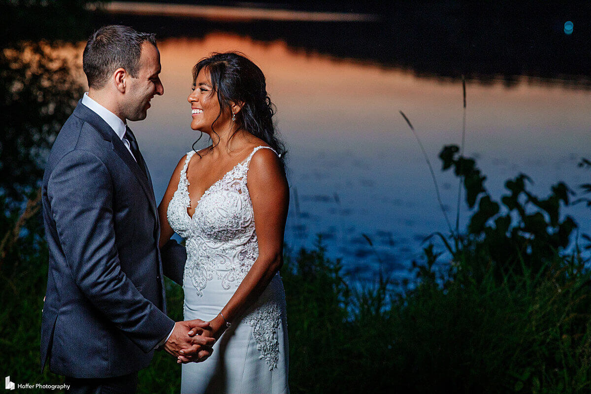 A photo from a gallery showing a bride and groom standing in front of a lake at sunset.