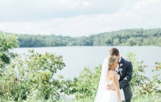 A bride and groom embrace outdoors in front of a lake at Lake House Inn, surrounded by greenery and trees—a picture-perfect scene for any wedding guest following A Wedding Guest Guide to Lake House Inn.