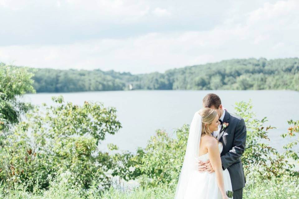 Lake-House-Inn-timeless-trends-header-image A bride and groom embrace outdoors in front of a lake at Lake House Inn, surrounded by greenery and trees—a picture-perfect scene for any wedding guest following A Wedding Guest Guide to Lake House Inn.