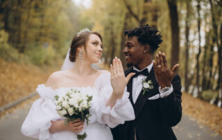 Bride and groom stand outdoors on a tree-lined path at the Lake House Inn, showing their wedding rings and smiling; the bride holds a white bouquet, capturing the joy of fusion weddings at this beautiful venue.