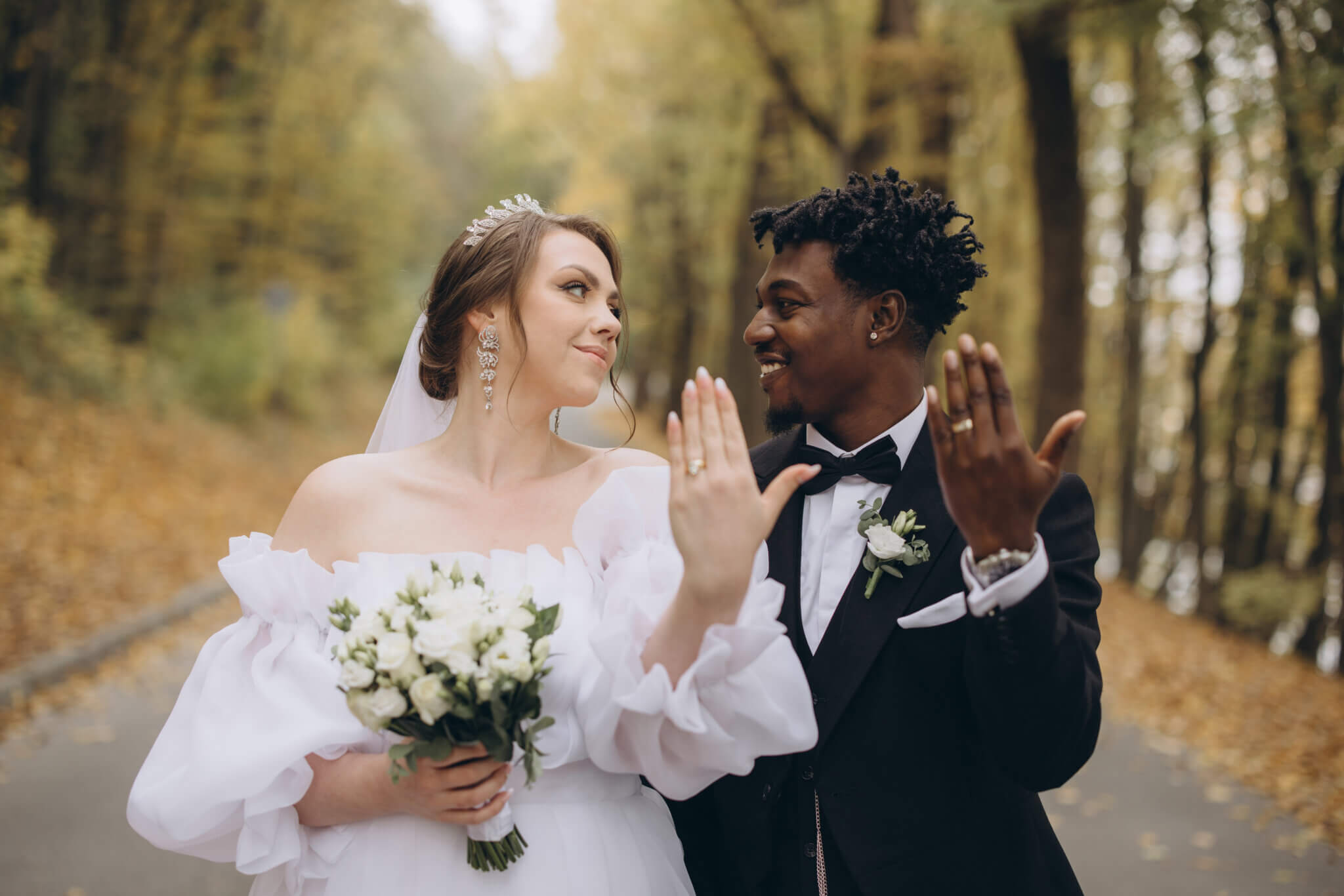 Bride and groom stand outdoors on a tree-lined path at the Lake House Inn, showing their wedding rings and smiling; the bride holds a white bouquet, capturing the joy of fusion weddings at this beautiful venue.